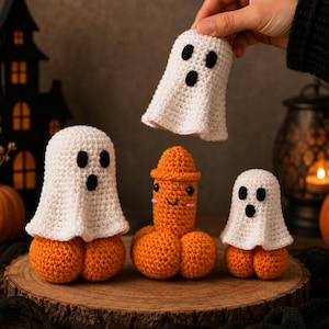 May include: Three crocheted figures on a wooden base. Two figures are white ghosts with black eyes and mouths sitting on orange pumpkin bases. One central figure is an orange phallic shape with a smiling face and pink cheeks on a pumpkin base.