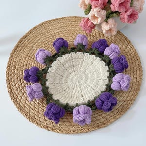 May include: A handmade crochet coaster featuring a central cream-colored circle surrounded by a ring of purple and lavender crochet flowers with green stems. The coaster sits on a woven, round placemat.