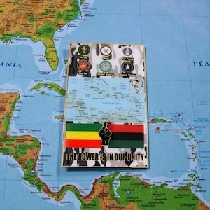 May include: A rectangular pin featuring a map of the Caribbean Sea, military branch emblems, and the text "THE POWER IS IN OUR UNITY." The pin has a gold border and a camouflage background.