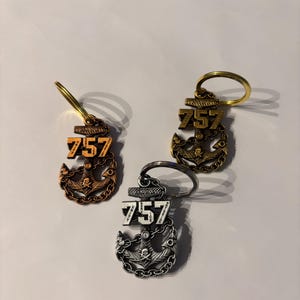 Puede incluir: Tres llaveros metálicos en bronce, dorado y plateado, cada uno con un diseño de ancla y el número "757" en la parte superior. Los llaveros tienen un anillo para sujetar las llaves.