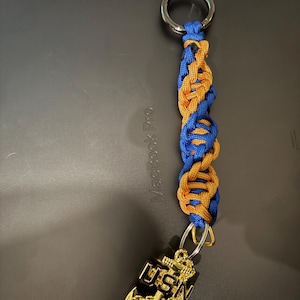 Puede incluir: Un llavero trenzado azul y dorado con un anillo plateado. El llavero presenta un dije de ancla de color dorado con las letras "USN". El llavero probablemente está hecho de paracord.