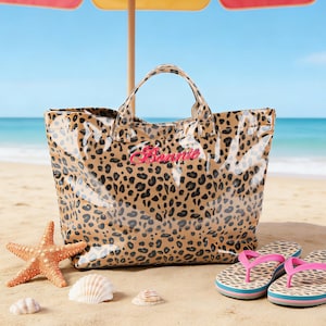 Könnte beinhalten: Strandszene mit einer Tragetasche mit Leopardenmuster, dem Namen "Barbie" in Rosa und passenden Flip-Flops. Ein Seestern und Muscheln liegen im Sand. Ein gestreifter Sonnenschirm spendet Schatten. Meer und Himmel sind im Hintergrund.