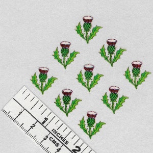 Pode incluir: Nove pequenos adesivos de cardo coloridos dispostos em padrão de diamante. Cada adesivo apresenta um caule e folhas verdes, uma cabeça de flor roxa e um fundo branco. Uma régua com polegadas e centímetros está no canto inferior esquerdo.