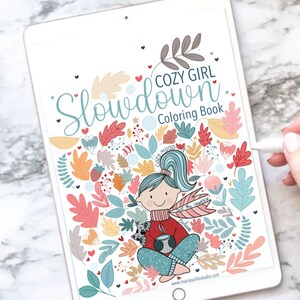 Op de afbeelding: Een digitale illustratie van een meisje met een kopje koffie en een klein dier dat in een cirkel van bladeren en harten zit. De tekst "Cozy Girl Slowdown Coloring Book" staat bovenaan de afbeelding.