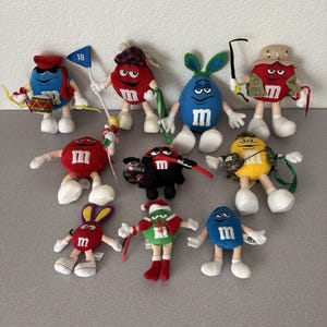 May include: A collection of ten M&M plush toys in various colors including red, blue, yellow, green, and black. The characters are dressed in diverse outfits such as a Santa suit, bunny ears, a fishing vest, a plaid hat, and a drum set accessory.
