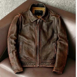 Könnte beinhalten: Eine dunkelbraune Lederjacke mit Used-Optik, ausgestattet mit einem klassischen Kragen, einem durchgehenden Frontreißverschluss und zwei seitlichen Reißverschlusstaschen. Die Jacke hat lange Ärmel mit verstärkten Details und ein beiges Innenfutter, das am Kragen sichtbar ist.