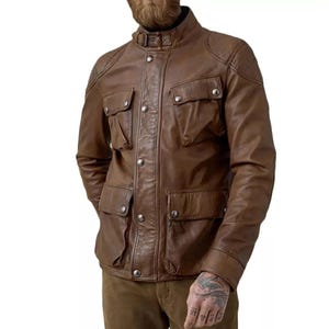 Könnte beinhalten: Eine braune Lederjacke mit Stehkragen und Schnallenverschluss, einer Knopfleiste mit silberfarbenen Knöpfen auf der Vorderseite sowie vier großen aufgesetzten Taschen. Die Jacke hat lange Ärmel und gesteppte Details an den Schulterpartien.