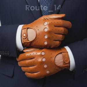 May include: Tan leather driving gloves with perforated details and a buttoned wrist strap. The gloves have a classic design and are worn over a white collared shirt and a navy blue suit. The text "Route_14" is visible in the background.