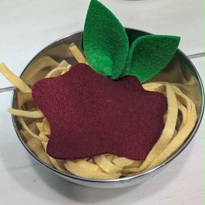 May include: A felt food toy in a stainless steel bowl. The toy features yellow felt noodles, a maroon felt meatball, and green felt basil leaves. The bowl is set against a white surface.