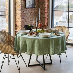 May include: A round dining table set for a meal, featuring a light green tablecloth and two wicker chairs. The table is set with white plates, bread, fruit, and candles. The table has a black metal base. The background includes a brick wall and a window.