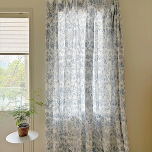 May include: White and blue floral patterned curtains hanging in front of a window. The curtains are made of a lightweight fabric and feature a detailed botanical design. The curtains are hung on a silver rail.