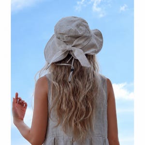 Peut inclure: Une femme porte un chapeau de soleil beige clair avec un grand nœud à l'arrière. Le chapeau est assorti à une robe sans manches de couleur similaire. L'arrière-plan est un ciel bleu vif avec quelques nuages.