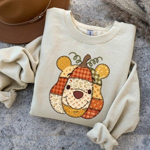 May include: A cream-colored sweatshirt featuring a pumpkin design with a bear face. The pumpkin is made of patchwork fabric in orange, yellow, and green. A brown hat is visible in the background. The sweatshirt is a fall-themed item.