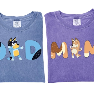 May include: Two folded t-shirts, one blue and one purple. The blue shirt has the word "DAD" with a cartoon dog character. The purple shirt has the word "MOM" with a cartoon dog character.