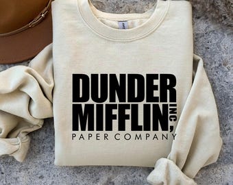 May include: A cream-colored sweatshirt with the text "Dunder Mifflin Paper Company" in bold black letters. The sweatshirt is folded, with the sleeves and collar visible. A brown hat is partially visible in the upper left corner.