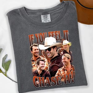 May include: Dark gray Comfort Colors t-shirt featuring a graphic of several men, one wearing a cowboy hat. The text "IF YOU FEEL IT! CHASE IT!" is printed in a distressed style. A brown hat is visible in the background.