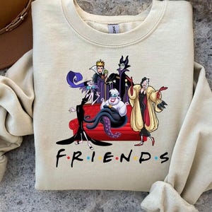 May include: Beige sweatshirt featuring a graphic of Disney villains sitting on a red couch. The characters include Ursula, the Evil Queen, Maleficent, and Cruella de Vil. The word "FRIENDS" is printed in black with colorful dots.