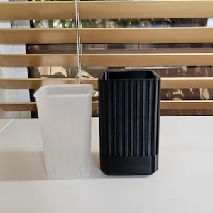 May include: Two planters sit side-by-side on a white surface. One is a translucent, square-shaped container with vertical ridges. The other is a black, square-shaped planter with vertical ridges. The background features a wooden window blind.