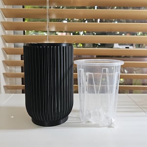 May include: A black, ribbed planter next to a clear plastic pot. The black planter has a cylindrical shape with vertical grooves. The clear pot has a rectangular shape with vertical supports. Both are on a white surface.
