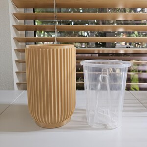 May include: A beige, ribbed, cylindrical planter next to a clear plastic pot with a built-in water level indicator. The planter has a rounded base and a vertical ribbed design. The clear pot is empty and has a handle.