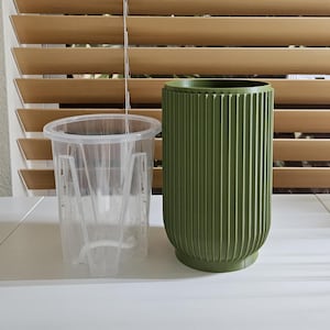 May include: A clear plastic plant pot with a handle is next to a green, ribbed plant pot. The green pot has a cylindrical shape and a rounded base. The background includes a window with wooden blinds.