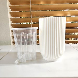 May include: A clear plastic plant pot with drainage holes sits next to a white, ribbed ceramic planter. The planters are on a white surface, with a wooden blind in the background.