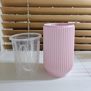 May include: A clear plastic plant pot insert next to a pink, ribbed, cylindrical planter. The planter has a rounded base and is designed to hold the insert. The background includes a white surface and wooden blinds.