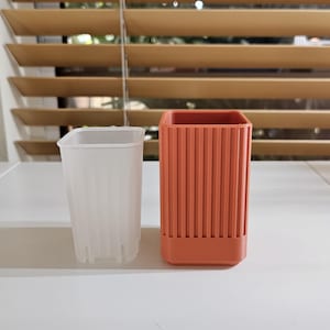 May include: Two planters sit side-by-side on a white surface. One is a translucent white square pot with vertical ridges. The other is a square, terracotta-colored pot with vertical ridges. The background features a window with wooden blinds.