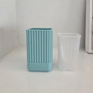 May include: A light blue rectangular container with vertical ribbed detailing on the exterior and a separate, translucent white inner liner. The container has a smooth, solid base and a square opening, designed for holding items upright.