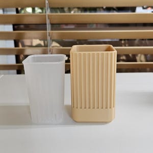 May include: Two square-shaped containers. One is a translucent, clear plastic cup with vertical ridges. The other is a beige, ribbed container. Both are sitting on a white surface, with a wooden blind in the background.