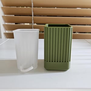 May include: A clear plastic plant pot with a white drainage string next to a green, ribbed, square-shaped planter. The planters are on a white surface, with a wooden blind in the background.