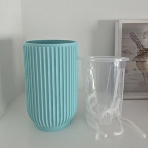 May include: A light blue cylindrical planter with a vertical ribbed texture and a separate clear plastic inner liner featuring integrated drainage slots and white cotton wicking cords extending from the base for self-watering functionality.
