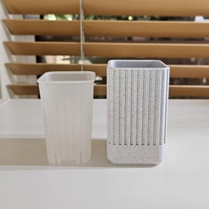 May include: Two square-shaped planters. One is a clear, ribbed plastic pot. The other is a speckled gray and white planter with vertical ridges. Both planters are empty and sit on a white surface.