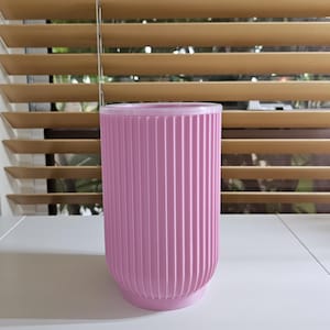 May include: A cylindrical light pink planter featuring a vertical ribbed texture along its exterior surface. The vessel has a smooth, rounded base and a flat, open top rim, presenting a consistent monochromatic color throughout the entire structure.