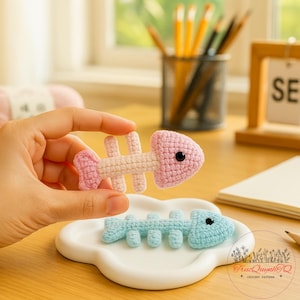 Peut inclure: Ornements en forme d'arêtes de poisson crochetées, roses et bleu clair. L'arête de poisson rose est tenue en l'air, tandis qu'une bleue claire repose sur un plat blanc en forme de nuage. Les ornements ont des yeux en boutons noirs.