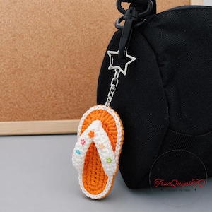 Könnte beinhalten: Ein gehäkelter orange-weißer Flip-Flop-Schlüsselanhänger mit bunten Sternakzenten. Der Schlüsselanhänger ist an einer schwarzen Tasche mit einem silbernen sternförmigen Verschluss befestigt. Das Flip-Flop-Design ist detailliert und handgefertigt.