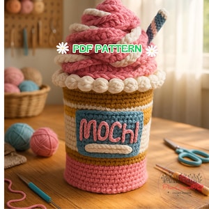 Puede incluir: Una bebida mochi de ganchillo hecha a mano, con una cobertura de crema batida rosa y blanca, una pajita a rayas y una etiqueta azul que dice "Mochi". La taza tiene una base rosa, un centro a rayas amarillas y blancas y un ribete de cuentas blancas. También se ve el texto "PDF PATTERN".
