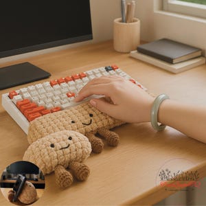 Puede incluir: Un reposamuñecas de teclado de ganchillo beige y un objeto más pequeño a juego con caras sonrientes. El teclado tiene teclas blancas y naranjas. Una mano descansa sobre el teclado. Una pulsera verde está en la muñeca de la persona.