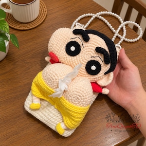 May include: A beige crochet tissue holder shaped like a cartoon character with thick black eyebrows, black hair, and red sleeves. The character wears yellow shorts and has a white pearl bead strap. A white tissue protrudes from the center of the figure.