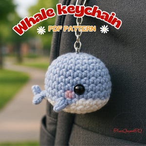 May include: A crocheted blue whale keychain with a white belly and pink cheeks. The whale has a black eye and small fins. The keychain is attached to a silver chain. Text on the image reads "Whale Keychain PDF Pattern".