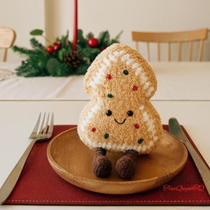 Puede incluir: Un muñeco de hombre de jengibre de peluche con una cara sonriente, detalles rojos y verdes y piernas de ganchillo marrones. El muñeco está sobre un plato de madera con un mantel individual rojo, tenedor y cuchillo. Decoración festiva de vacaciones en el fondo.