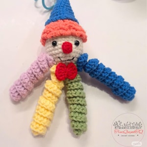 May include: A colourful, handmade crochet clown doll. It features a white face, red nose, and bow tie, with a blue hat and orange and pink trim. The arms and legs are in shades of pink, yellow, green, and blue. A whimsical, handcrafted item.