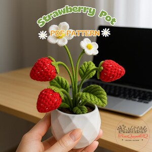 Op de afbeelding: Een handgemaakte gehaakte aardbeienpot met rode aardbeien, witte bloemen en groene bladeren. De pot is wit en geometrisch. De tekst "Strawberry Pot PDF Pattern" is in de afbeelding.