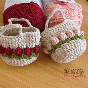 May include: Two small, handmade crochet baskets with handles, one decorated with red flowers and green leaves, the other with pink flowers and green leaves. The baskets are beige, and the background includes balls of yarn.