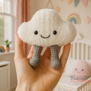 May include: A soft, white cloud-shaped toy with black button eyes and a smiling mouth. It has grey crocheted legs. The toy is held in a hand, with a fluffy texture, ideal for nursery decoration. The toy is made of soft material.