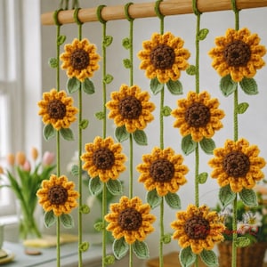 Puede incluir: Una cortina de ganchillo hecha a mano con un patrón repetido de girasoles. Los girasoles tienen pétalos amarillos y centros marrones, con hojas y tallos verdes. La cortina está suspendida de una barra de madera.