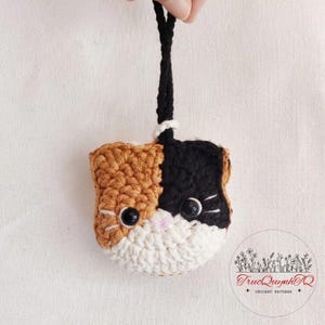 May include: A small, handmade crochet cat head ornament featuring a tri-color pattern of tan, black, and white yarn. The cat has two round black plastic eyes, a small pink stitched nose, and white whiskers. A black yarn loop is attached to the top.