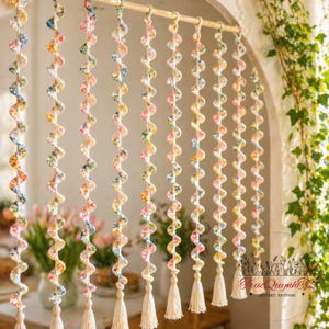 Op de afbeelding: Een decoratief gordijn gemaakt van gehaakte spiraalslingers met bloempatronen in pastelkleuren. Elke streng is versierd met een kwast aan de onderkant. Het gordijn hangt aan een houten roede en creëert een grillig en artistiek element voor de woondecoratie.