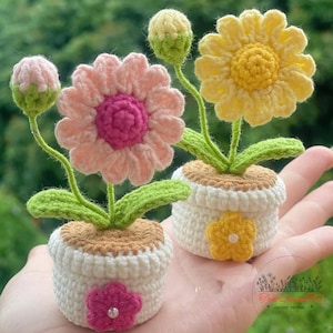 Könnte beinhalten: Zwei gehäkelte Blumenarrangements in kleinen weißen Töpfen. Eines hat eine rosa Blume mit einem magentafarbenen Zentrum, das andere eine gelbe Blume. Beide haben grüne Stiele und Blätter. Die Töpfe sind mit kleinen Blumen verziert.