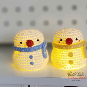 Könnte beinhalten: Zwei gehäkelte Schneemann-Lampen mit leuchtendem Innenraum. Jeder Schneemann hat eine rote Nase, schwarze Augen und einen Schal. Ein Schal ist blau, der andere gelb. Die Lampen stehen auf einer weißen Oberfläche.